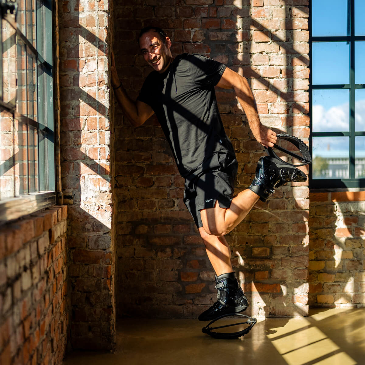 Hombre apoyado en una pared de ladrillo, posando y haciendo estiramientos mientras lleva puestas unas botas de rebote Fit Boots en interiores.