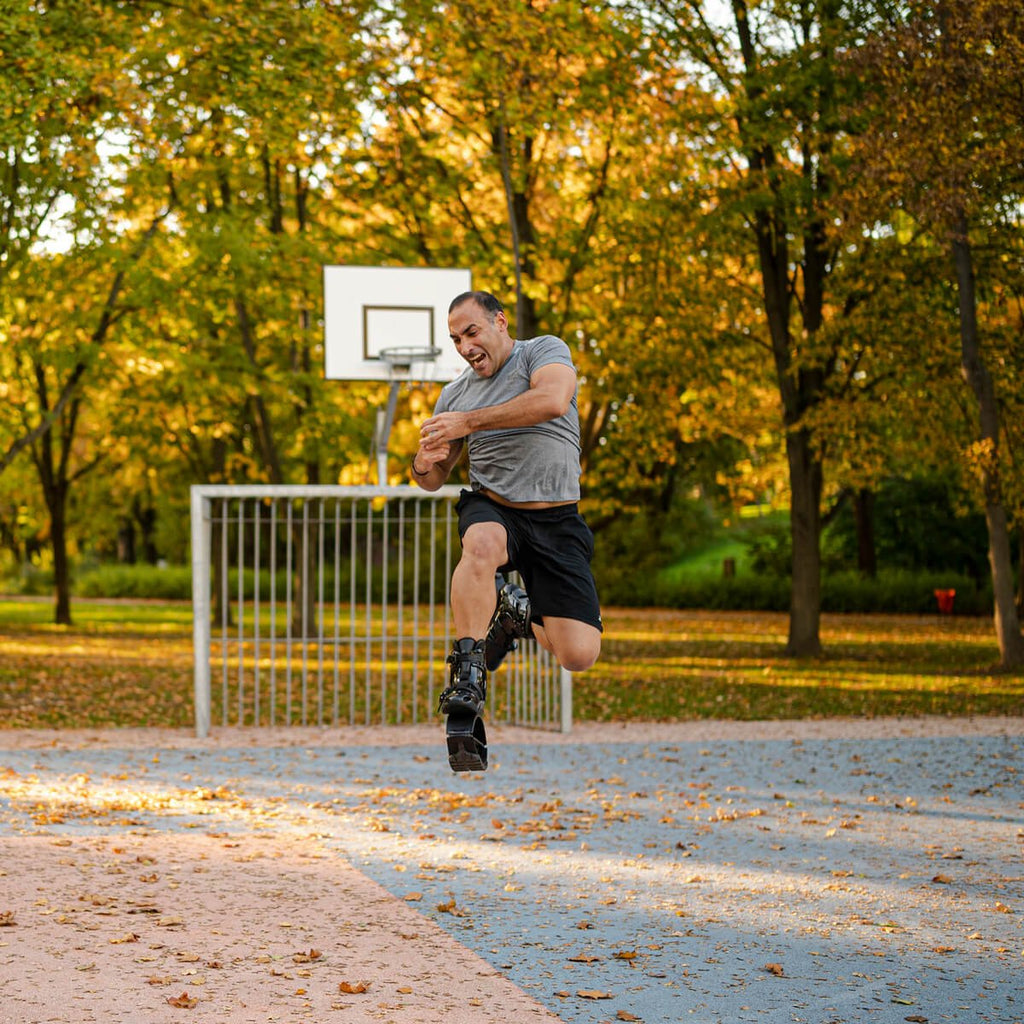 Hombre saltando en el aire mientras lleva puestas unas botas de rebote Fit Boots en una cancha de baloncesto al aire libre rodeada de árboles con follaje otoñal.