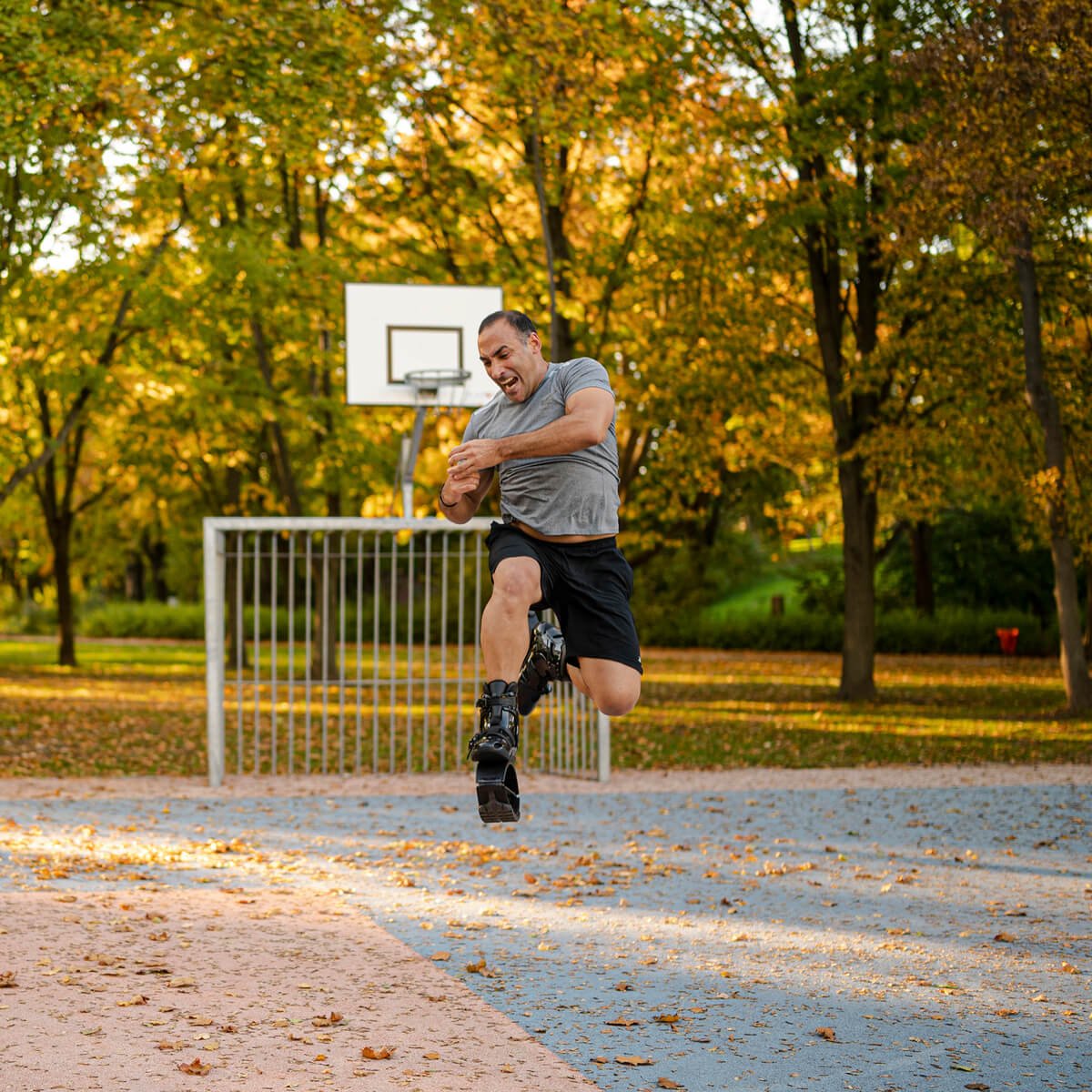 Hombre saltando en el aire mientras lleva puestas unas botas de rebote Fit Boots en una cancha de baloncesto al aire libre rodeada de árboles con follaje otoñal.